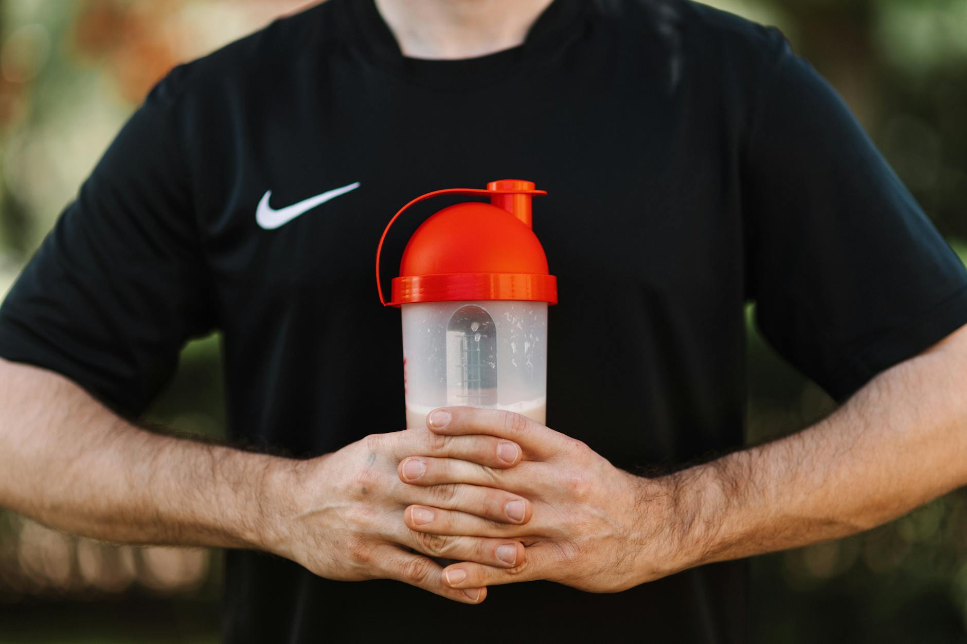 Adult man holds a protein shake in a red lid tumbler outdoors, wearing a black shirt.