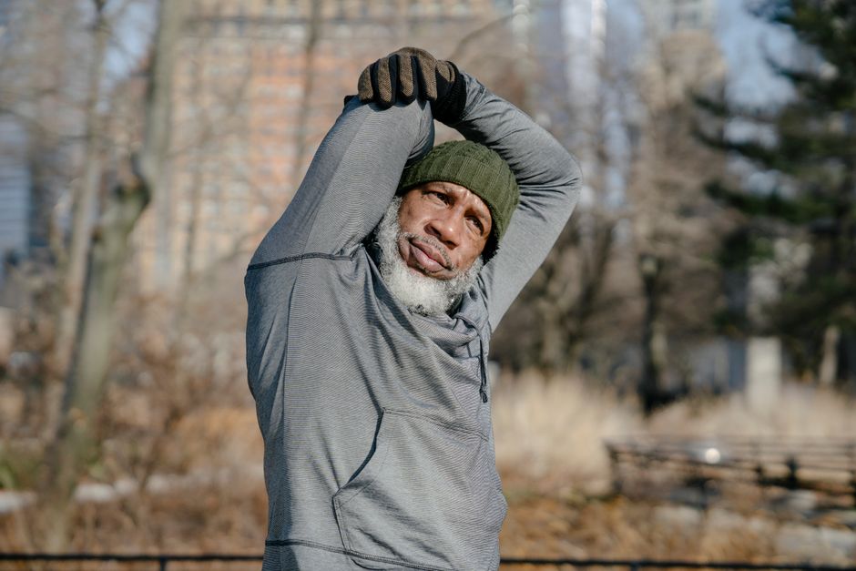 African American elderly bearded man in hoodie and cap looking away and raising arms during warm up in autumn park