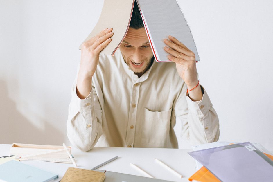 A stressed adult male worker sits at a desk with open notebooks, exhibiting signs of frustration and burnout.